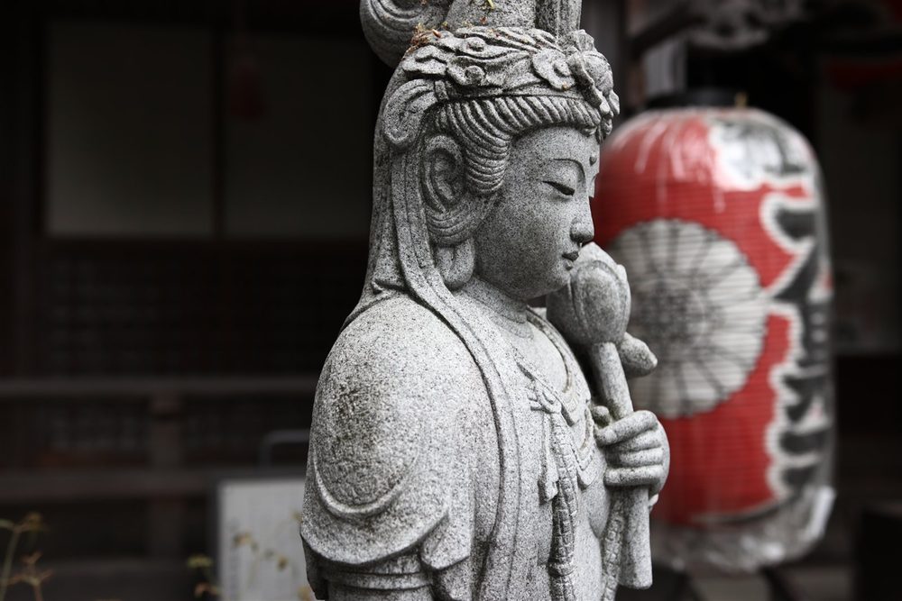 A granite statue of Kannon holding a lotus bud, beside a red paper lantern at a temple