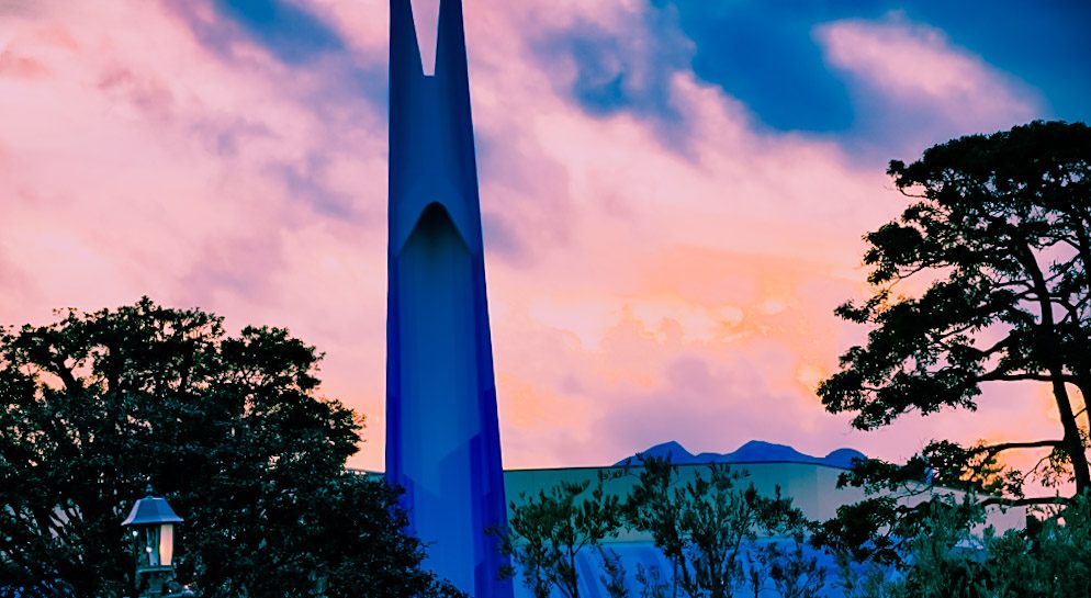A tall blue spire silhouetted against a pink and orange sunset sky, with dark trees and an old-style lamp post