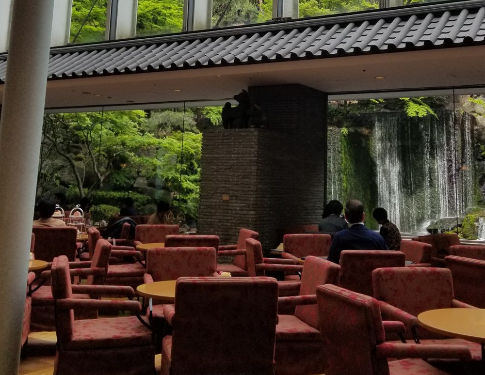 A tall glass atrium lounge with red armchairs, looking out to green garden trees and a tall waterfall