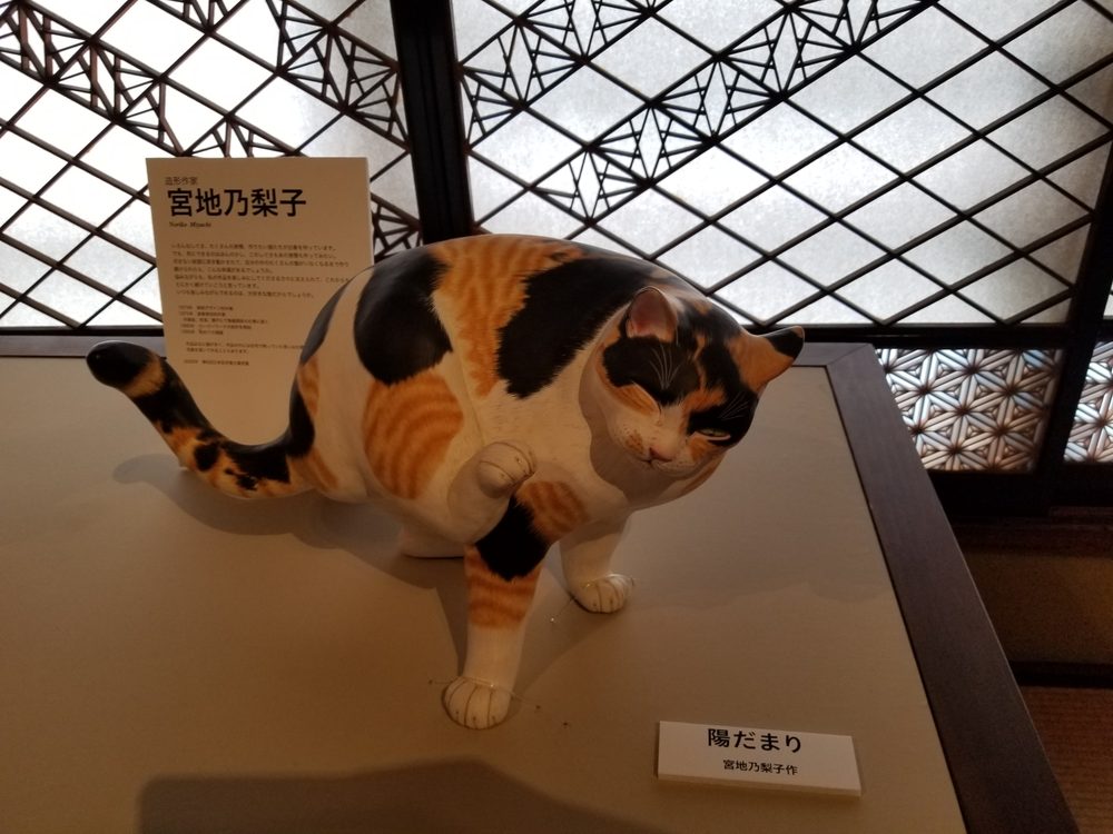 A life-size sculpture of a calico cat stretching on a low plinth, beneath a geometric wooden lattice