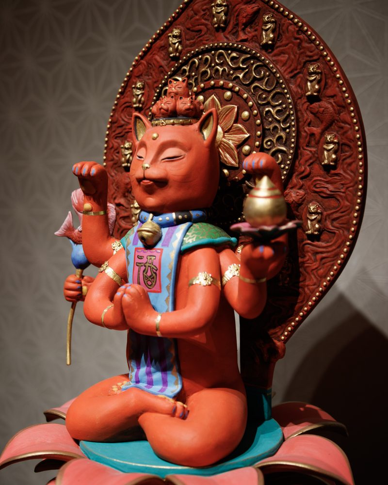 A red cat-faced Buddha figure with several arms, seated on a lotus in front of a carved halo
