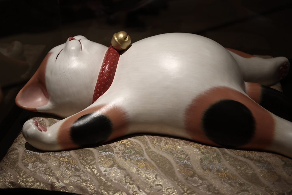 A white ceramic calico cat with a red collar and gold bell, lying on its back on a brocade cushion