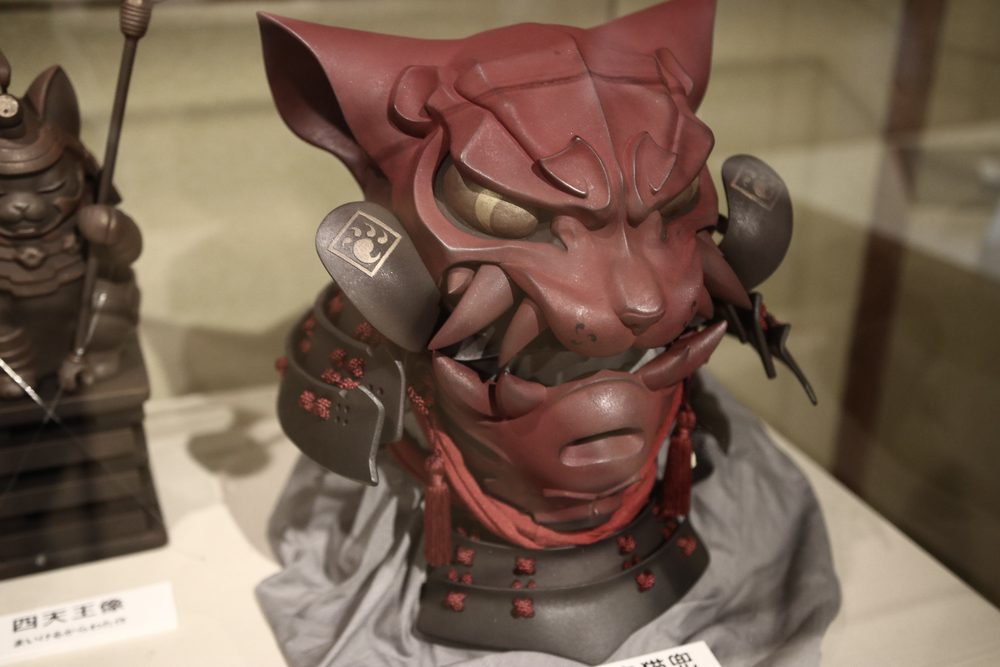 A sculpted cat's head wearing red samurai armor and a helmet, in a glass display case