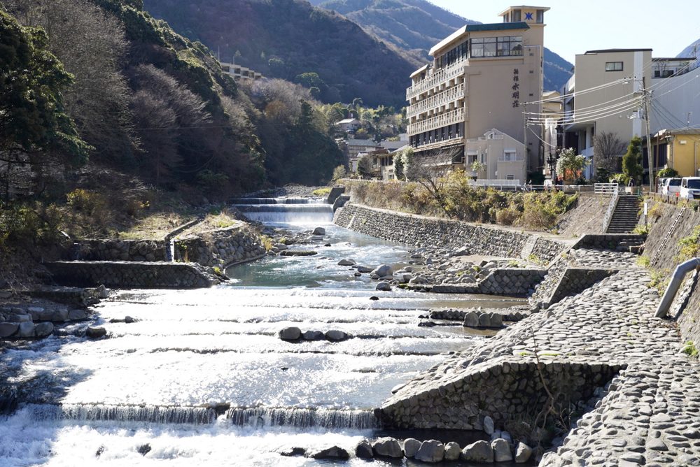 A clear mountain river tumbling over low stone weirs beside a hot-spring hotel and wooded hills in Hakone