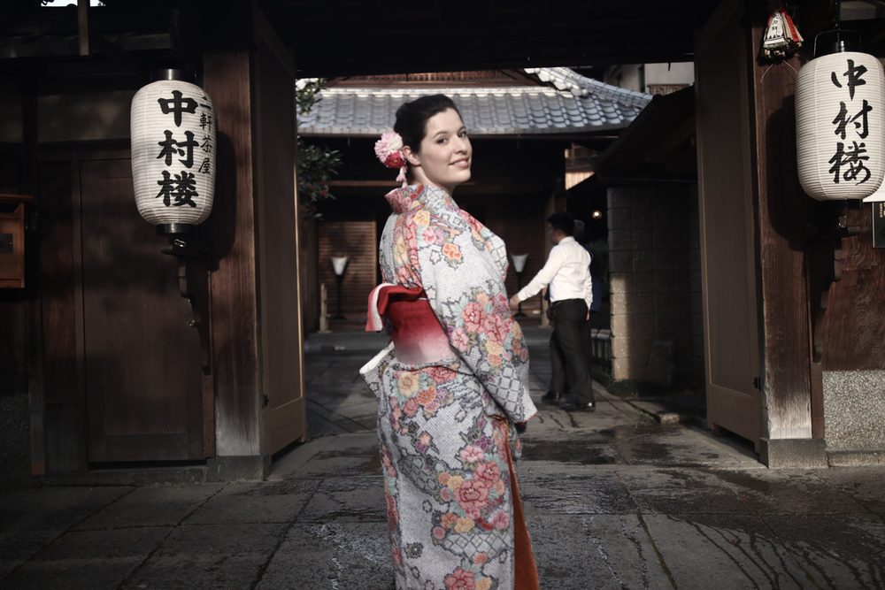 Joanna in a rented floral kimono looking back over her shoulder at a wooden gate hung with white paper lanterns in Kyoto