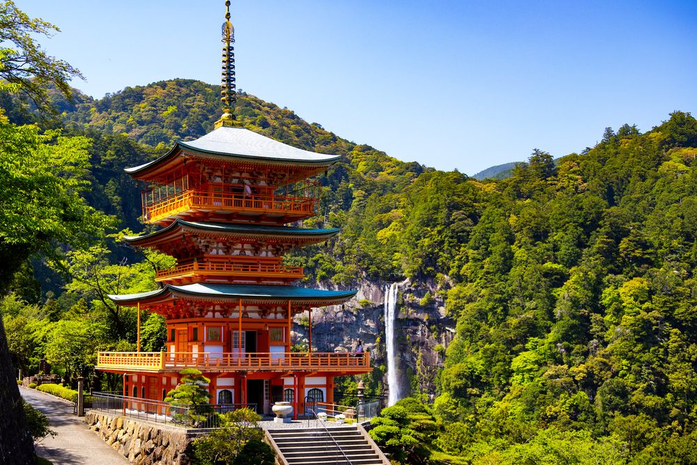 The vermilion three-story pagoda of Seiganto-ji in front of Nachi Falls and forested green mountains under a clear sky