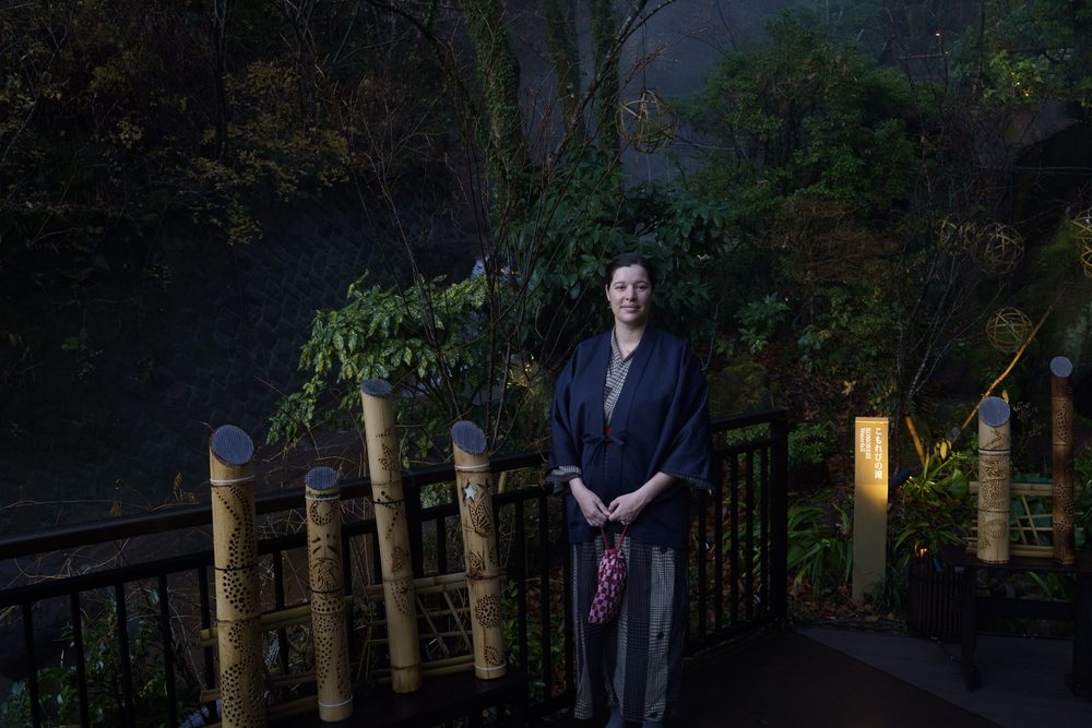 Joanna in an indigo yukata standing by a railing with carved bamboo lanterns, in a garden at dusk