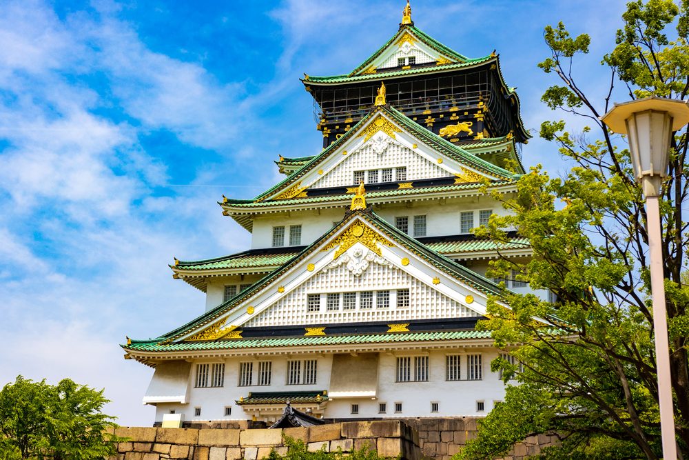 Osaka Castle's white and green main tower with gold trim, against a blue sky with a tree and a lamp post in the foreground