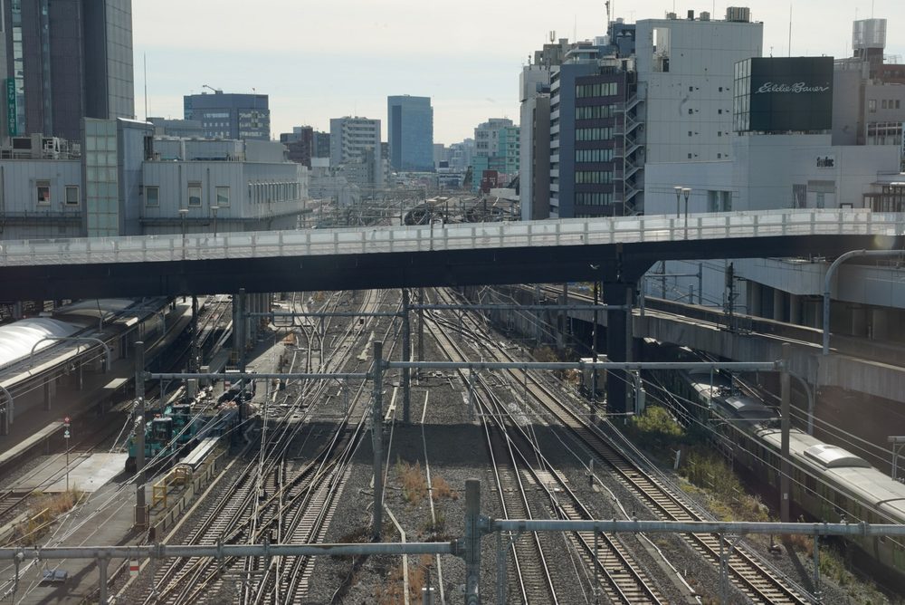 Many parallel railway tracks and overhead wires seen from an overpass, with a train and city buildings beyond