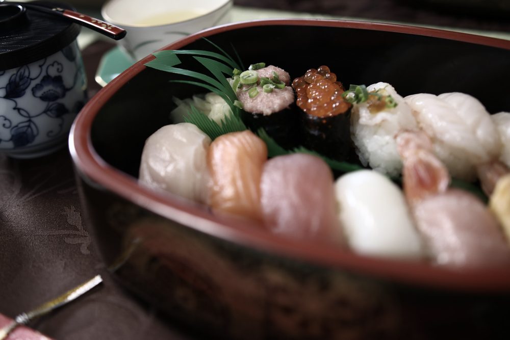 A lacquered box of nigiri sushi with salmon roe, scallop and shrimp, next to a blue and white teacup