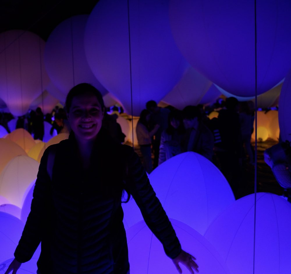 Joanna smiling among large glowing purple and amber balloon-like spheres in a dark teamLab room, with other visitors as dark silhouettes