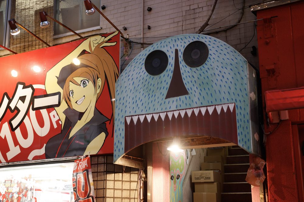 A blue monster-face awning over a small shop entrance beside a red arcade sign on a Tokyo side street