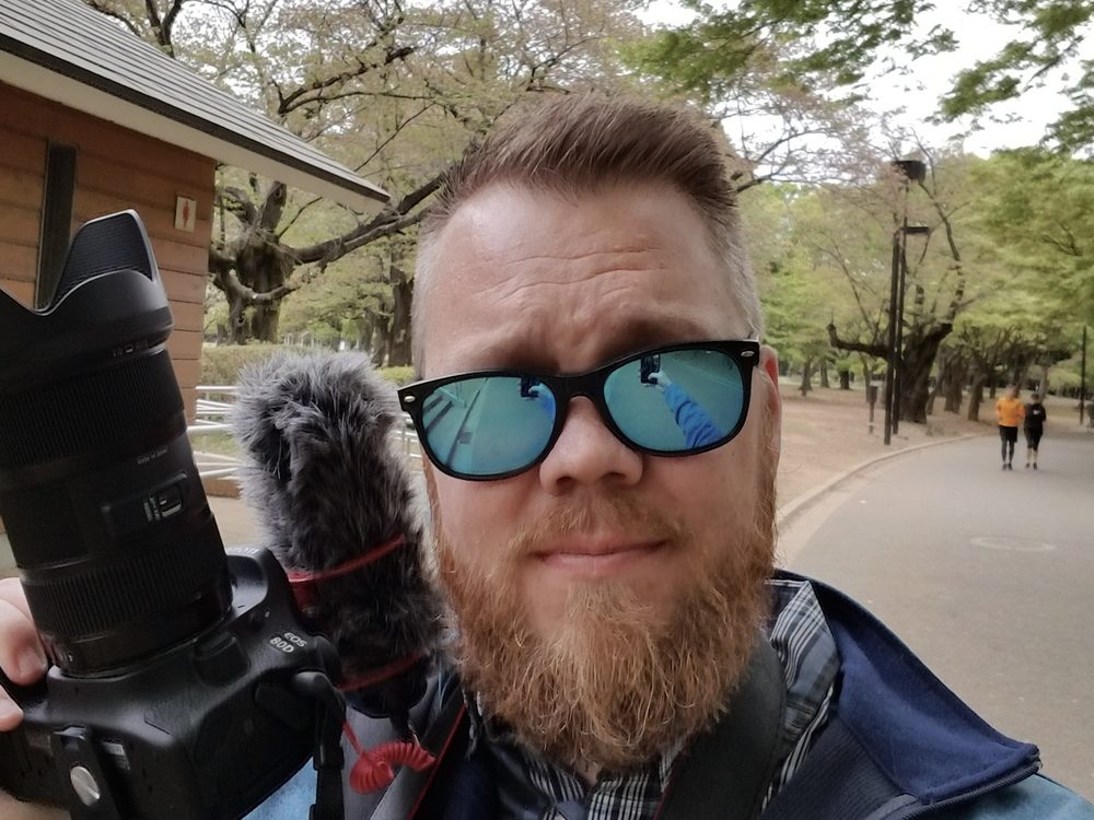 Travis in sunglasses holding a Canon camera with a furry microphone windscreen, on a tree-lined park path in Tokyo